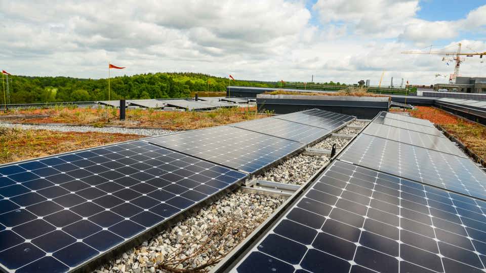 Svarta solceller som löper över ett tak, med grön skog till vänster och blå himmel med moln.