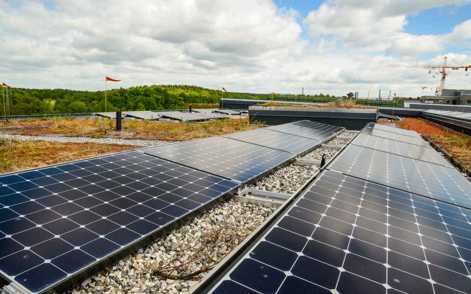 Svarta solceller som löper över ett tak, med grön skog till vänster och blå himmel med moln.